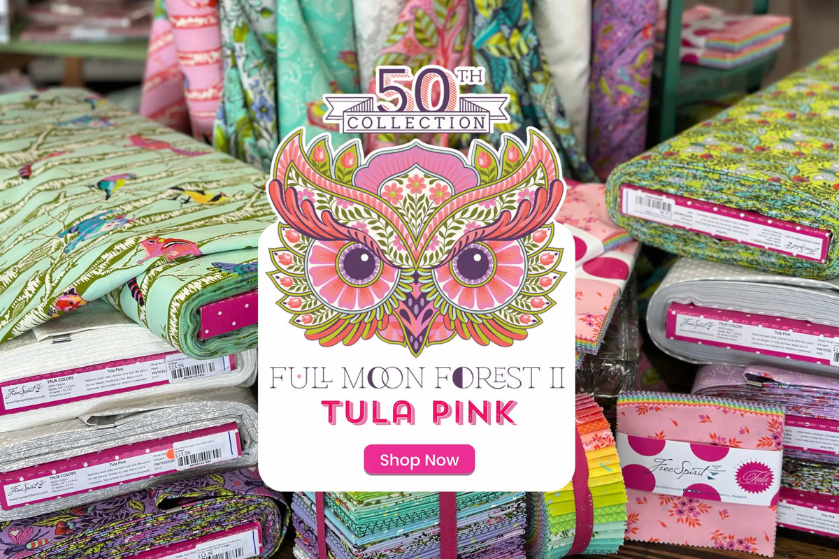 Stacks of colorful patterned fabric bolts and bundles with a sign displaying a decorative owl, reading “50th Collection, Full Moon Forest II, Tula Pink, Shop Now.”.