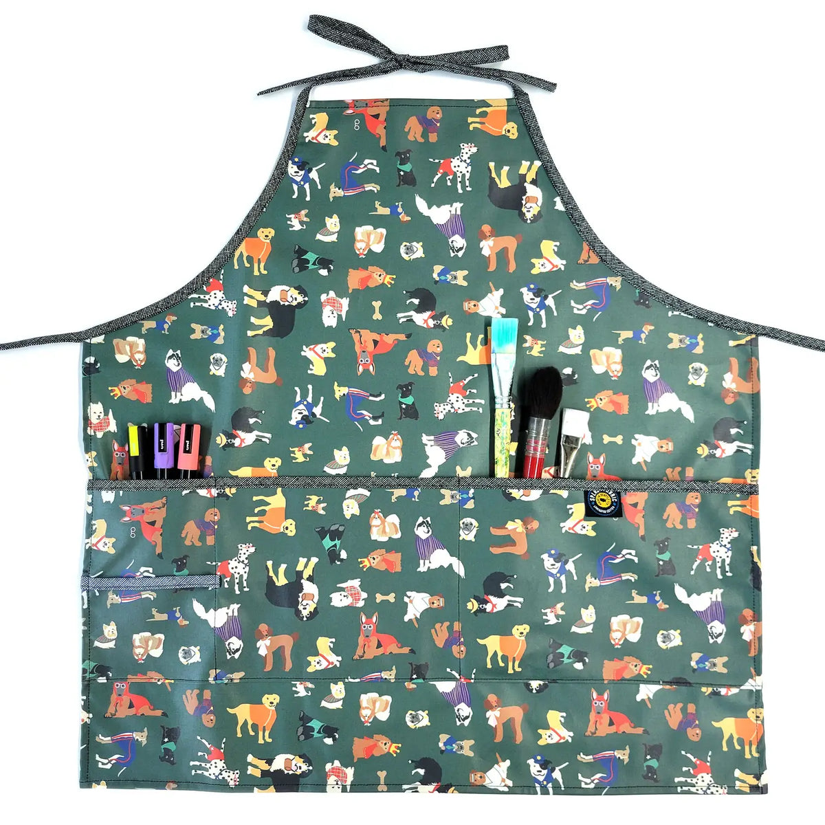 A green apron with cartoon dog illustrations, featuring three front pockets holding pens, markers, and paintbrushes. The apron has neck and waist straps for tying.