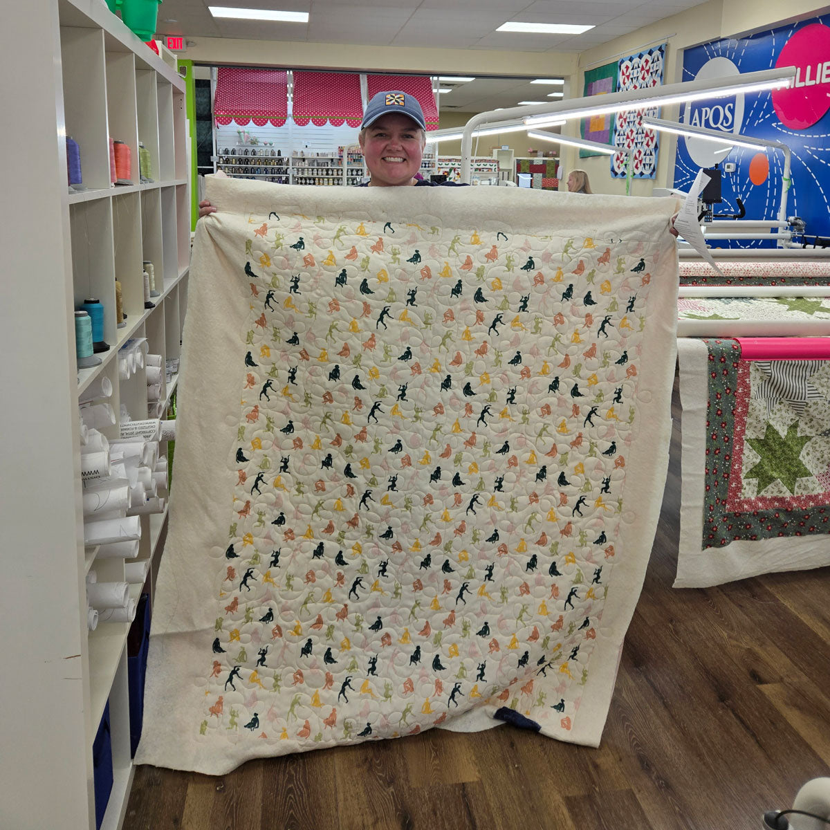 A person in a baseball cap smiles while holding up a handmade quilt inside a craft or sewing store. The quilt features a repeating pattern of colorful birds on white fabric. Shelves of supplies and other quilts are visible in the background.