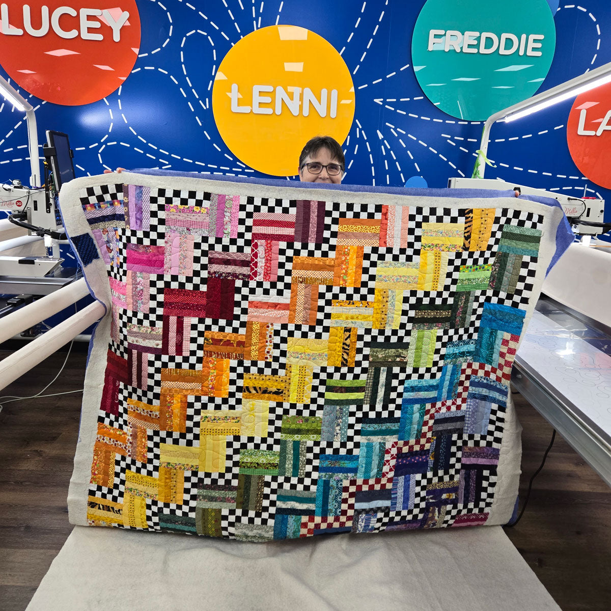 A person holds up a colorful patchwork quilt with a zigzag pattern, featuring vibrant blocks in various colors and a black-and-white border. Behind them, a blue wall displays large circles labeled Lucey, Lenni, Freddie, and Larry.