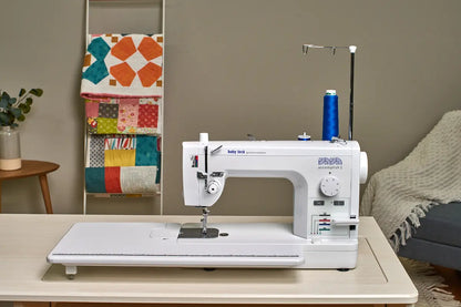 A Baby Lock Accomplish 2 Sewing Machine with FREE online sewing classes, threaded in blue, sits on a table. Behind it are colorful quilts on a ladder, a comfy chair draped with a blanket, and greenery arranged in a vase.