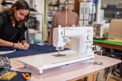 A Baby Lock Accomplish 2 Sewing Machine with FREE online sewing classes sits on a table with fabric, scissors, and tools. A woman works in the background, focused on measuring dark blue fabric, while shelves and sewing supplies fill the workspace.