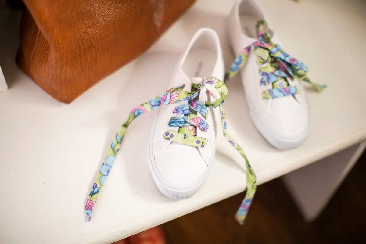 A pair of white sneakers with colorful floral-patterned fabric shoelaces, crafted using the Baby Lock Accomplish 2 Sewing Machine with FREE online sewing classes, sits on a white surface next to a brown leather bag.