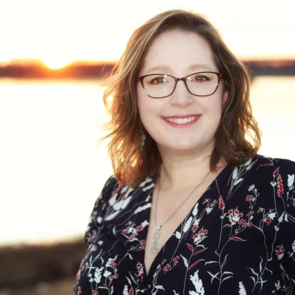 A woman with shoulder-length wavy hair and glasses smiles at the camera. She is wearing a dark floral blouse, standing outdoors near water with the sun setting in the background.