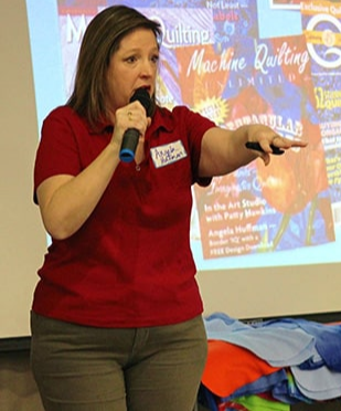 A woman in a red shirt holds a microphone and gestures while speaking in front of a colorful quilt-themed presentation screen.