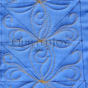 Blue fabric with intricate yellow-gold thread quilting in a symmetrical, swirling floral and geometric pattern. The quilting creates a decorative and textured design on the fabric’s surface.