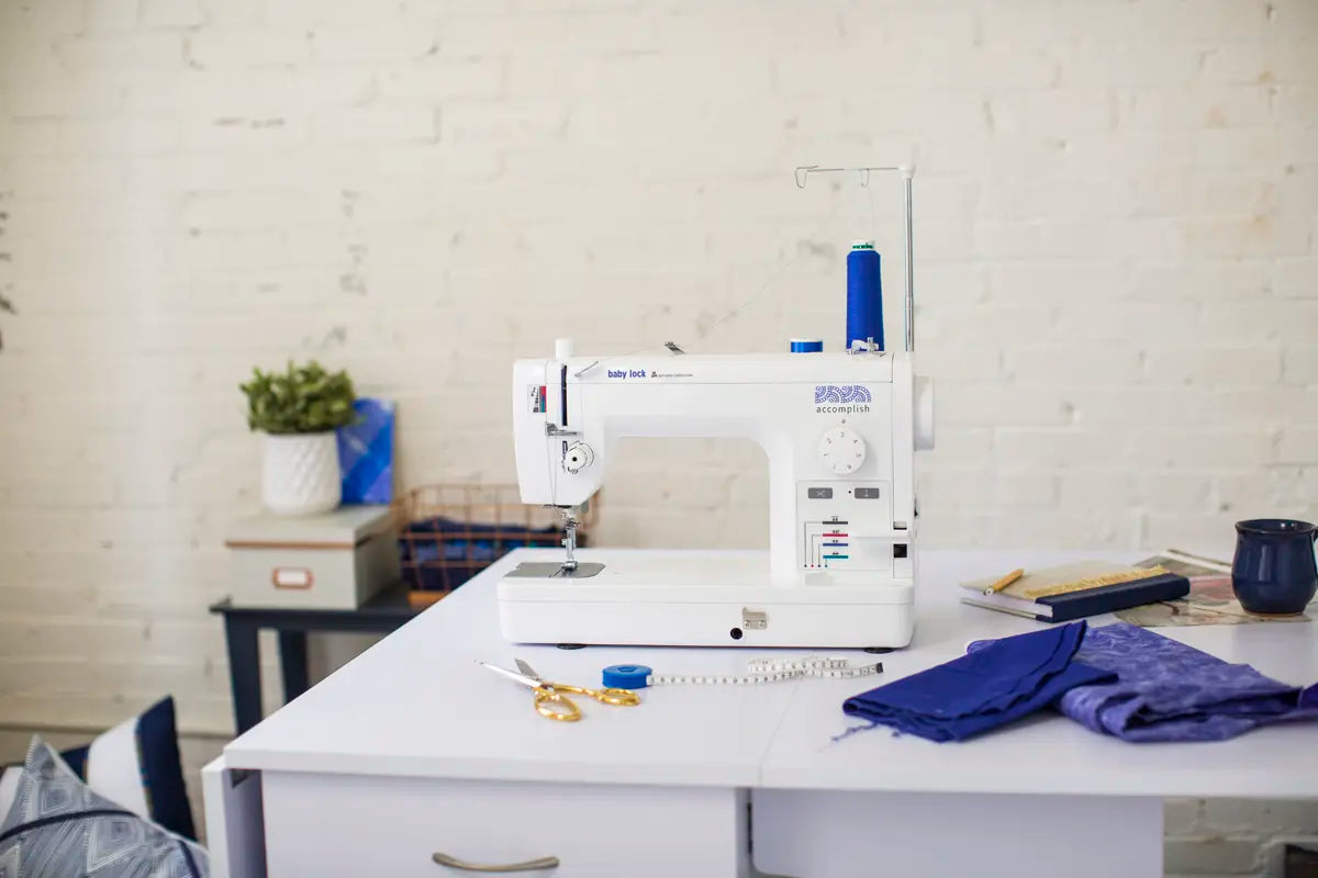 A Baby Lock Accomplish 2 Sewing Machine with FREE online sewing classes sits on a desk surrounded by fabric, scissors, measuring tape, and thread in a bright room with a white brick wall and a small plant.