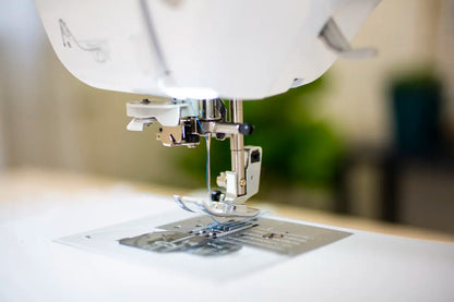 Close-up view of the Baby Lock Jubilant Sewing Machine needle and presser foot, with a thin blue thread through the needle, poised above the metal needle plate. The softly blurred background highlights the machine’s detail.
