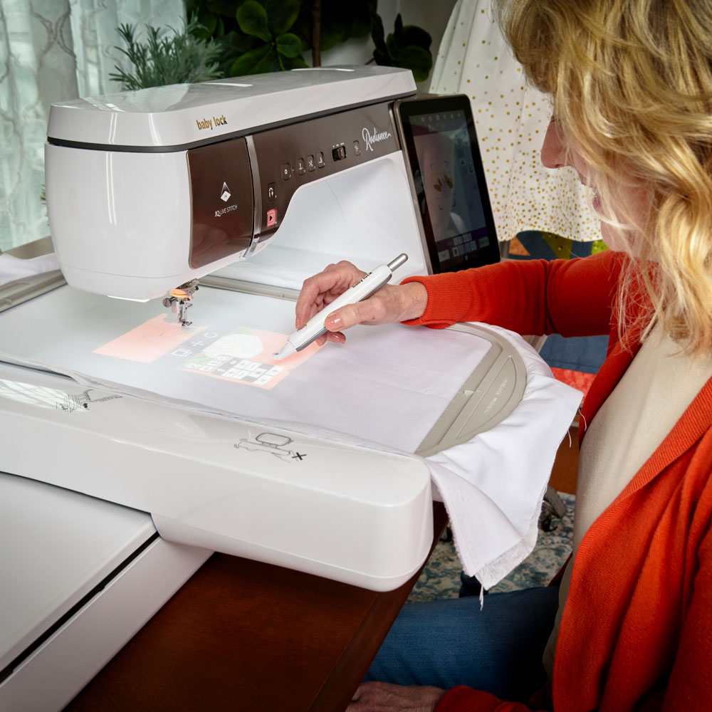 A woman uses a stylus to design embroidery on a touchscreen sewing machine, with white fabric placed under the machine’s needle and colorful patterns displayed on the screen.