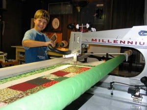 A smiling child uses a large quilting machine to sew a colorful patchwork quilt, guiding the fabric under the machines needle in a well-lit workspace.
