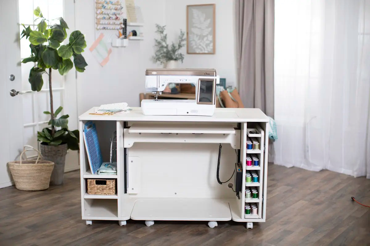 A white sewing table with the Baby Lock Chorus Sewing Machine, thread spools, and organized craft supplies sits in a bright room with wood floors, plants, and large windows draped with sheer curtains.