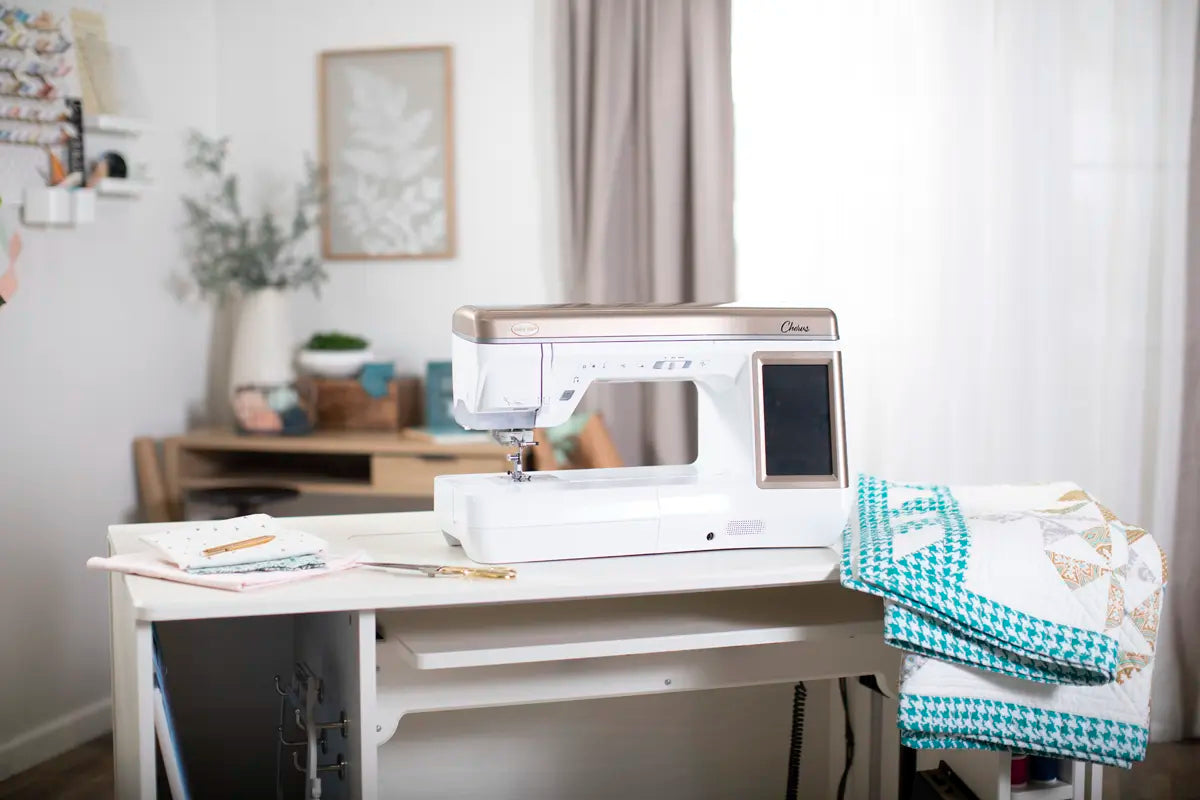 A Baby Lock Chorus Sewing Machine by Baby Lock rests on a white table with folded quilts and supplies. The softly lit, cozy room—decorated and furnished with a desk—is perfect for projects using its Compact Digital Dual-Feed System.