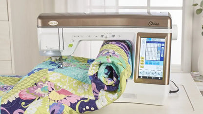 The Baby Lock Chorus Sewing Machine-FLOOR MODEL, featuring a digital touchscreen and dual-feed system, is stitching a colorful quilt of green, purple, blue, and pink patterned fabrics by a window.