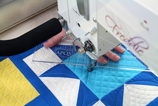 A person uses a longarm quilting machine to sew blue, white, and yellow geometric quilt blocks, guiding the fabric with a clear ruler for precise stitching.