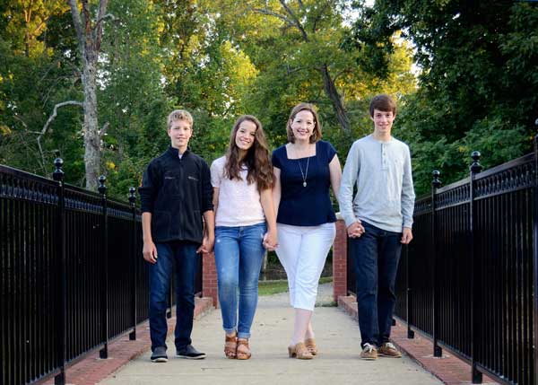 Four people, two adults and two teenagers, stand side by side holding hands on a pathway outdoors, flanked by black railings, with green trees in the background. They are all smiling at the camera.