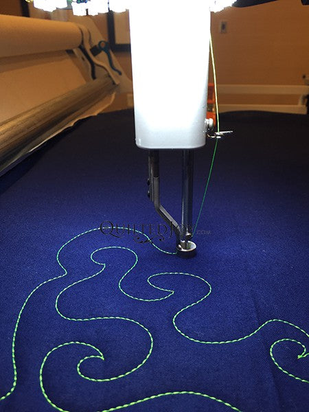 A sewing machine is stitching a wavy green pattern onto dark blue fabric, using a long arm quilting setup.