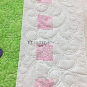 A section of a quilt featuring a green fabric with polka dots on the left and a white strip with pink squares and intricate quilted swirl patterns on the right. The word quiltedjoy.live is faintly visible on the white fabric.