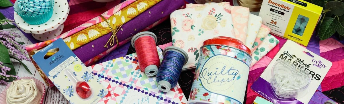 A colorful assortment of quilting supplies, including patterned fabric, spools of thread, quilt clips, needles, bobbins, quilting pins, and a decorative tin, all arranged on a quilted surface.