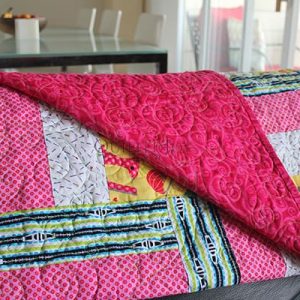 A colorful patchwork quilt with a bright pink, patterned underside is draped over a chair, with a dining table and chairs visible in the background.