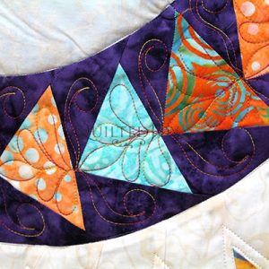 A close-up of a colorful quilt featuring a curved strip of orange and blue triangle patterns with swirly stitching on a dark purple background. The surrounding fabric is a light, marbled color.
