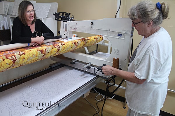 Two women work together on a quilting machine; one operates the machine stitching a floral design, while the other observes and speaks, possibly giving instructions or advice. Fabric with yellow and orange patterns is on the machine.