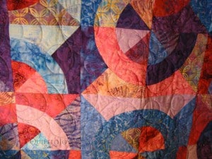 A colorful quilt featuring geometric patterns with overlapping circles and triangles in shades of red, pink, blue, and purple. The fabric has intricate, textured designs and stitching.