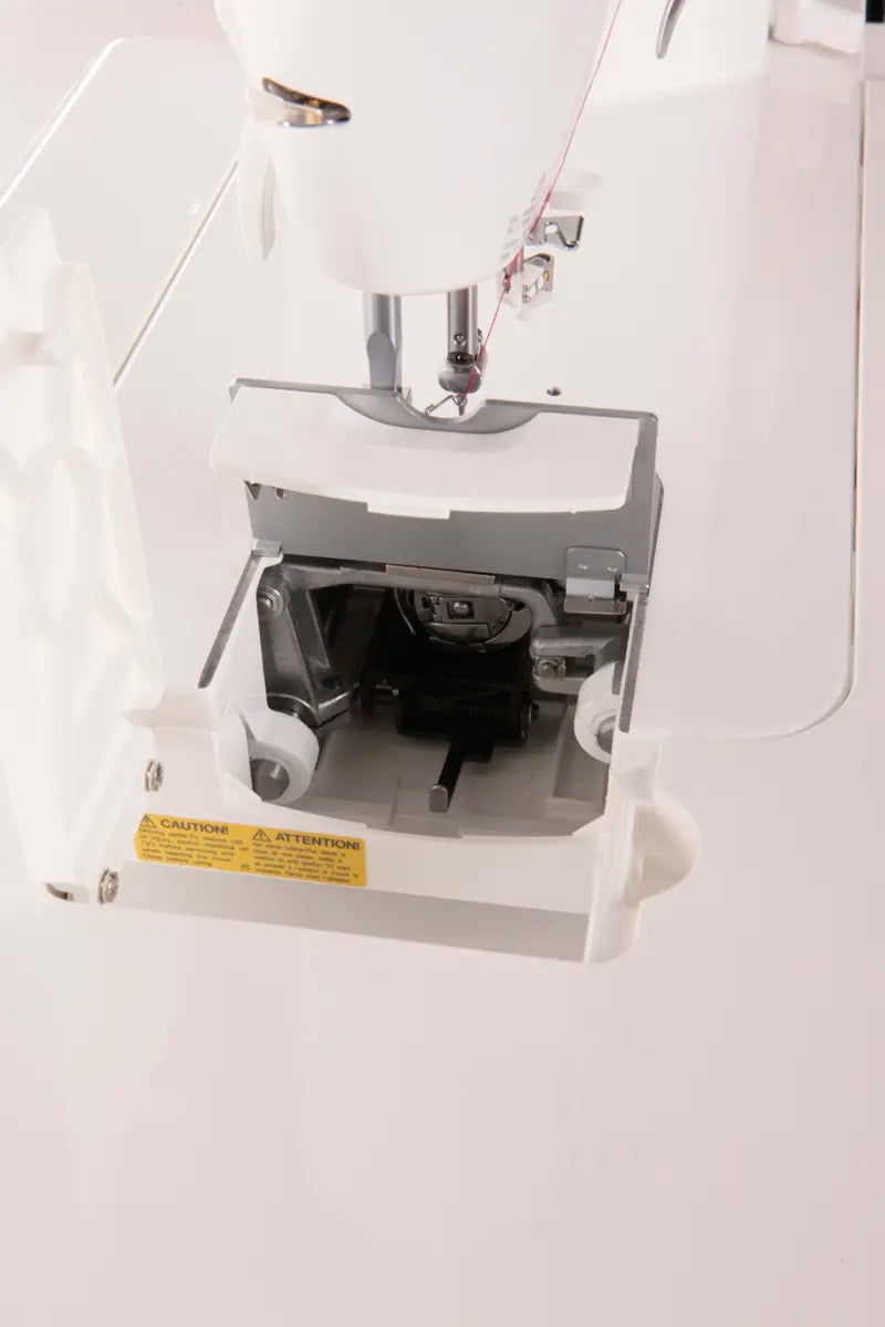 Close-up of an open JUKI TL-2000Qi Sewing and Quilting Machine bobbin case, displaying the internal compartment and a yellow caution label on the door.