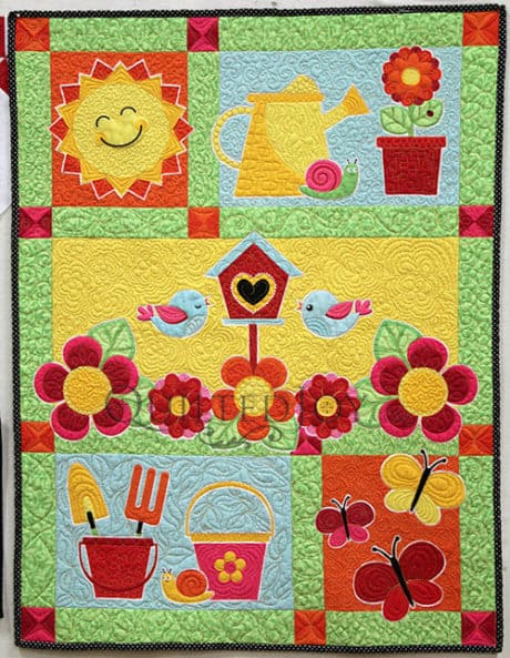 A colorful quilt featuring cheerful images: a smiling sun, watering can, flowerpots, birds, birdhouse, flowers, garden tools in a bucket, butterflies, and a basket, all on bright, patterned squares.