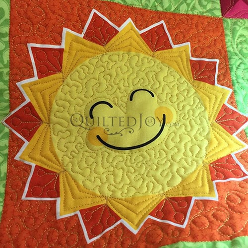 A colorful quilt square featuring a smiling sun with a yellow face, orange and red rays, and intricate stitching details on a green and orange patterned background.