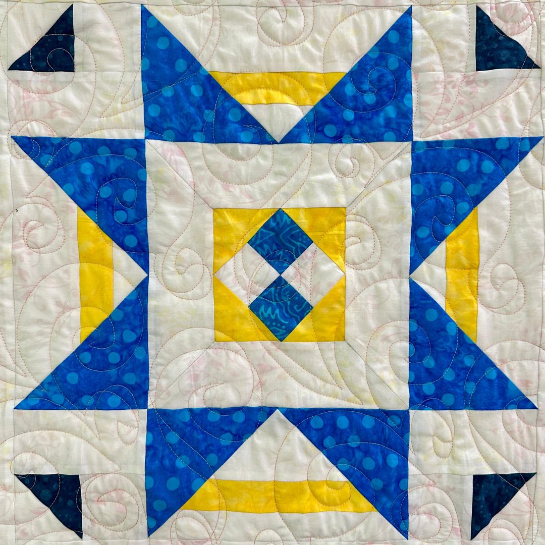 A quilt block featuring a geometric pattern with blue, yellow, and white fabrics. The design includes large blue triangles forming an eight-pointed star, with a smaller yellow and blue diamond pattern in the center and intricate quilting stitches.