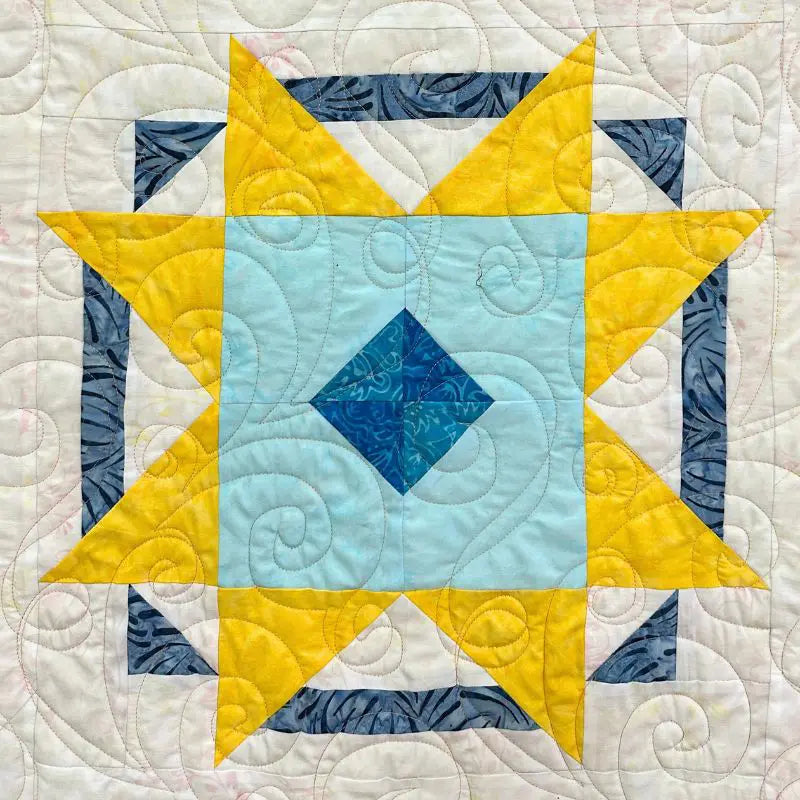 A colorful quilt block featuring a large yellow star, a light blue center with a blue diamond, dark blue corners, and a cream background with swirling quilted stitching patterns.