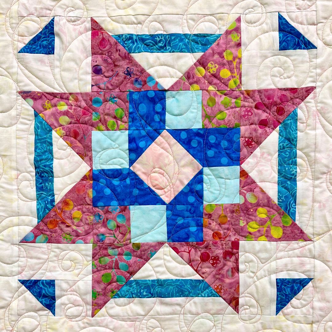 A colorful quilt block with a geometric star pattern, featuring blue, pink, and green floral fabrics, set against a light background with decorative quilted swirls.