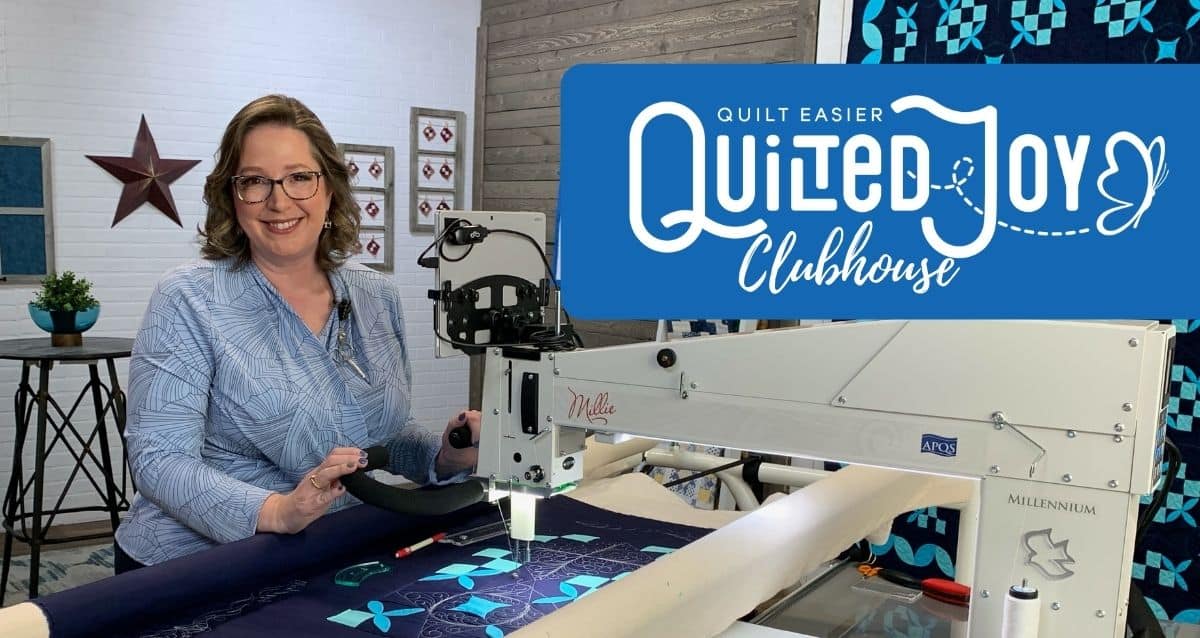 A woman smiles while using a quilting machine on a blue quilt in a bright studio. Behind her, a sign reads Quilted Joy Clubhouse with quilting graphics and a butterfly. Quilted art decorates the background.