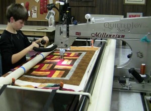 A person operates a quilting machine, guiding fabric with a geometric pattern through the machine in a workshop setting. The quilt has red, yellow, brown, and white designs.