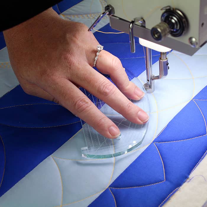 A close-up of a hand guiding a quilting ruler under a sewing machine needle while stitching a blue and light blue quilt with curved patterns. The person is wearing a ring.
