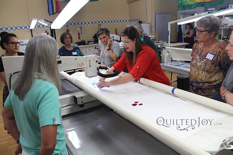 A group of women watch as an instructor in a red shirt demonstrates quilting techniques on a large quilting machine in a classroom setting. The scene includes fabric, sewing tools, and the Quilted Joy logo.