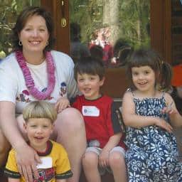 A woman wearing a pink lei sits outdoors with three young children, all smiling at the camera. The children wear colorful clothes and name tags. Trees and a glass door are visible in the background.