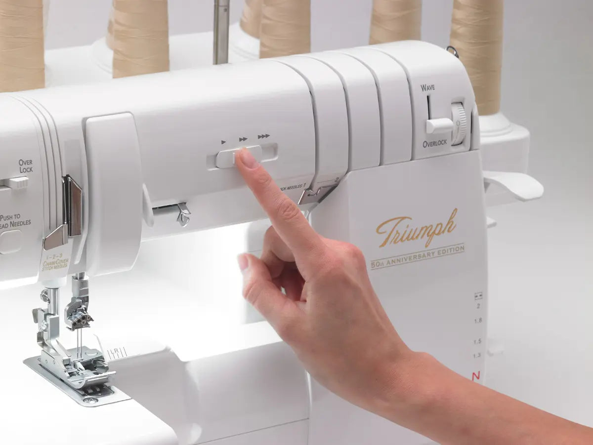 A hand presses a button on the Baby Lock Triumph Serger Machine with RevolutionAir threading, while several spools of beige thread sit in the background.