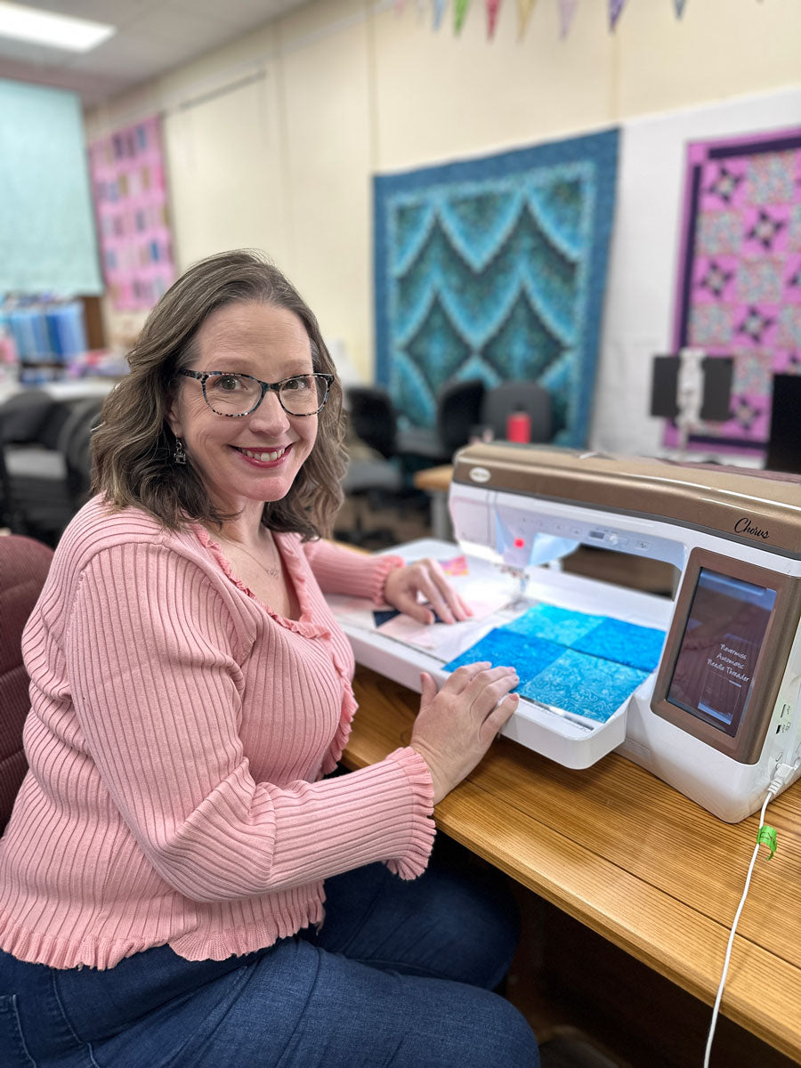A woman wearing glasses and a pink sweater smiles while working at a sewing machine with blue fabric in a room decorated with colorful quilts on the walls.