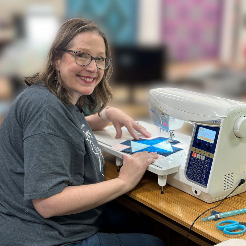 A woman with glasses smiles while using a sewing machine at a wooden table. She is working on a piece of blue and white fabric, with scissors nearby. The background is blurred.