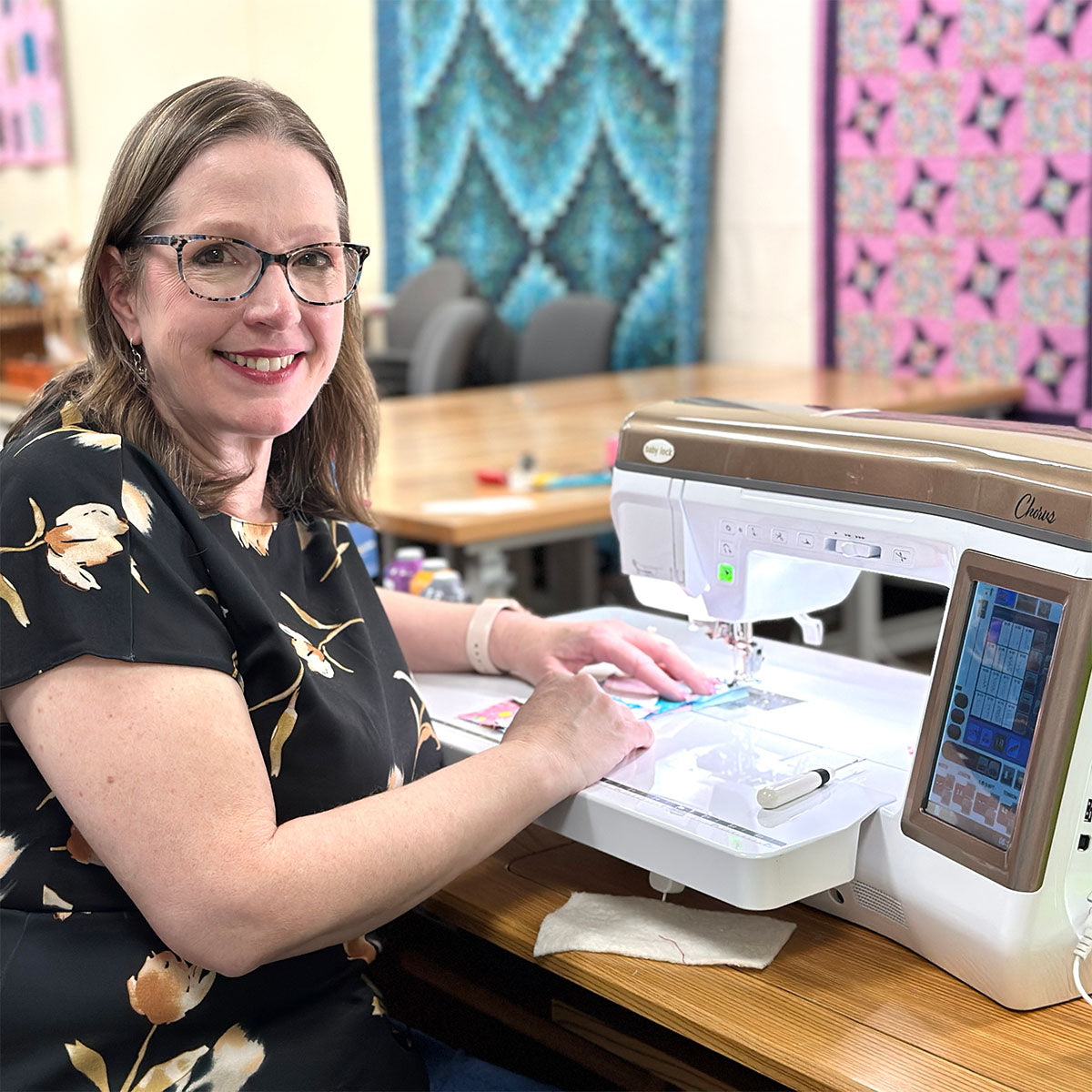 A woman wearing glasses and a black floral blouse sits at a table using a sewing machine, smiling at the camera. Quilted fabric designs hang on the walls in the background.