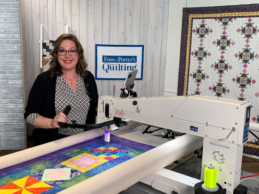 A woman stands smiling beside a long-arm quilting machine working on a colorful quilt. Behind her are finished quilts displayed on the walls, and a sign that reads Fons & Porters Quilting.