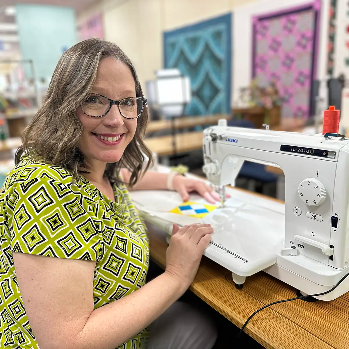 A woman with glasses and shoulder-length hair smiles while using a sewing machine to quilt in a colorful, well-lit workspace with patterned quilts hanging on the wall behind her.