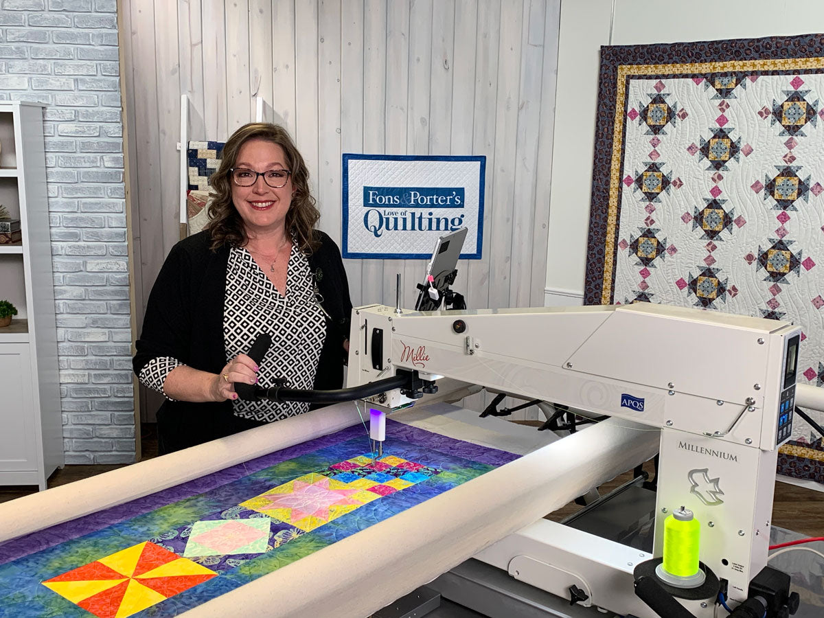 A woman stands beside a long-arm quilting machine working on a colorful quilt. Behind her is a sign that reads Fons & Porters Quilting, and a completed quilt hangs on the wall in a well-lit studio.