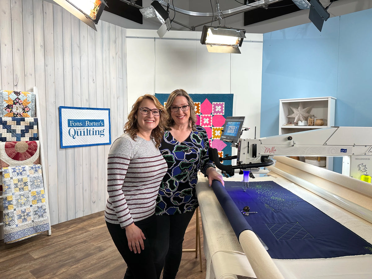 Two women smile and stand beside a long-arm quilting machine in a bright studio, surrounded by colorful quilted wall hangings and bright studio lights. A sign reads “Fons & Porter’s Quilting.”.
