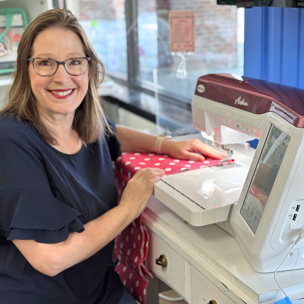 A woman wearing glasses and a navy blue top smiles while using a sewing machine to stitch red polka-dot fabric. She is seated at a white table in a brightly lit room.