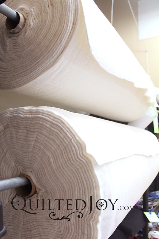Two large rolls of cream-colored quilt batting are stacked horizontally on metal rods. The thick, layered material is displayed in a well-lit store. The text quiltedjoy.live appears on the image.