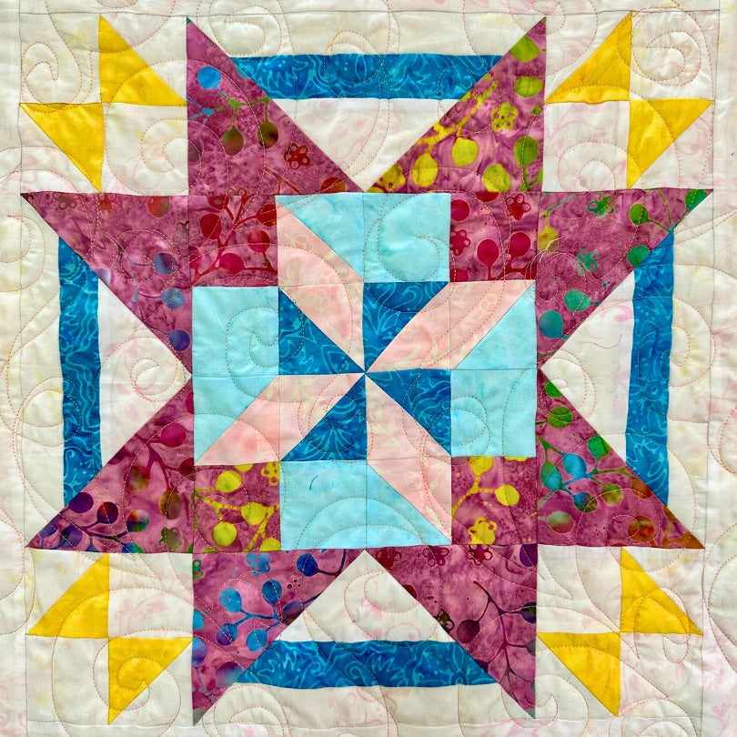 A colorful quilt block featuring a central blue and pink pinwheel, surrounded by purple, blue, green, and yellow geometric shapes, with intricate stitched patterns on a white background.