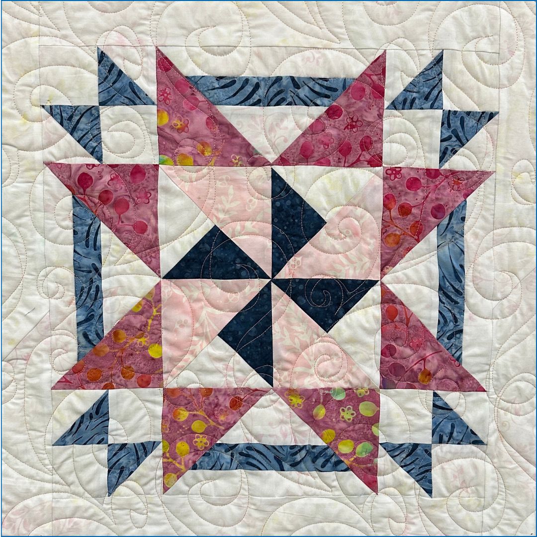 A colorful quilt block with a symmetrical geometric pattern in blue, pink, and white fabrics, accented with yellow dots and green flower prints, and intricate swirling quilt stitching throughout.