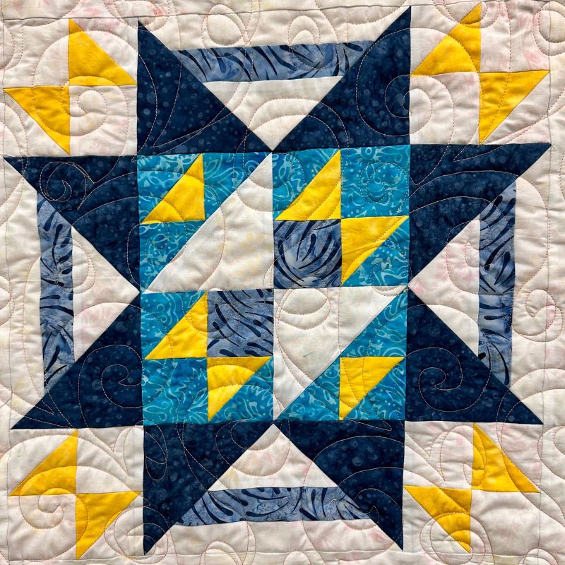 A colorful quilt square with a geometric pattern featuring blue, teal, yellow, and white fabrics. The design includes triangles, squares, and intricate stitching with swirling quilted lines across the surface.